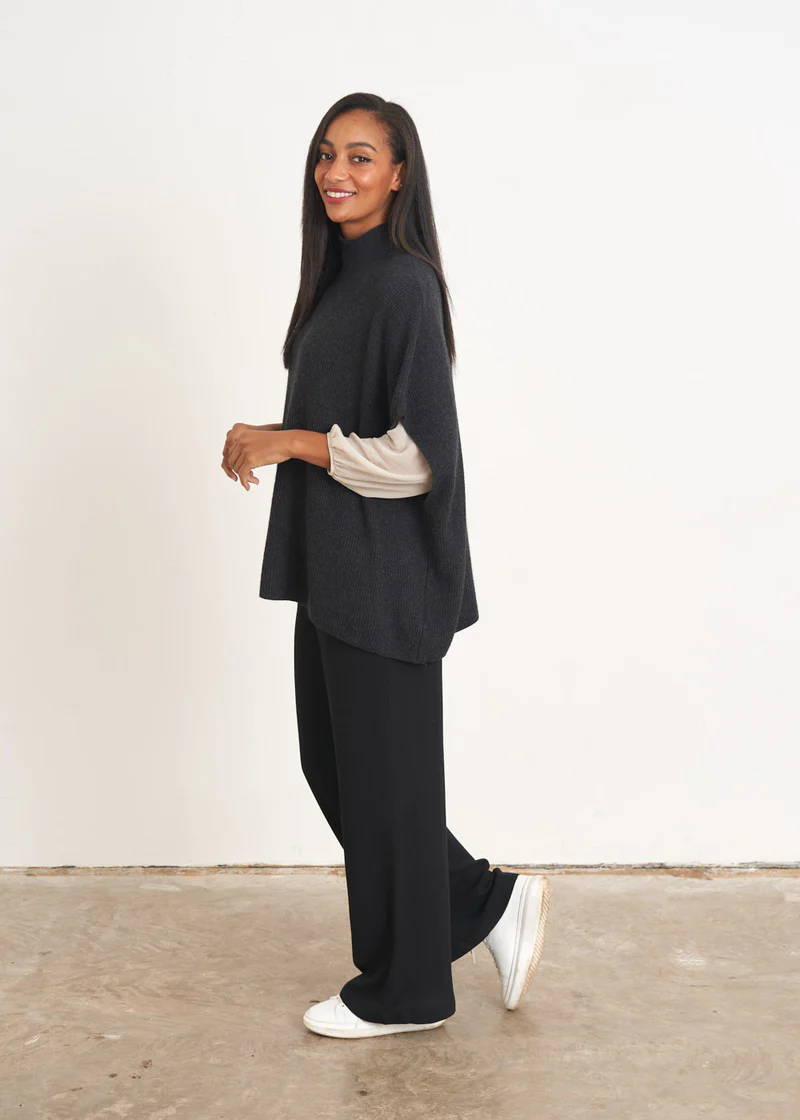 A model wearing a dark grey oversized sleeveless knitted sweater over a white top, black trousers and white trainers