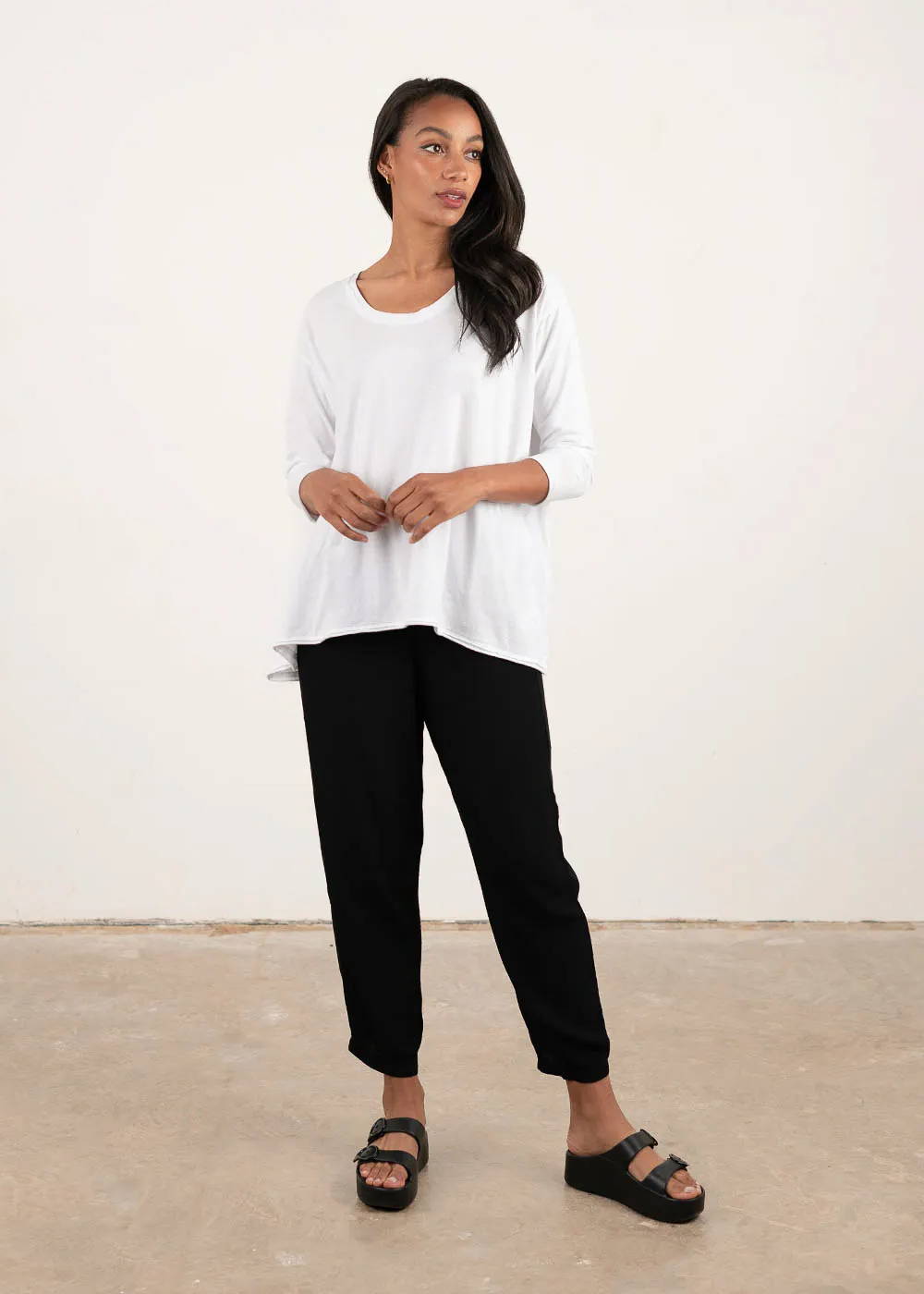 A model wearing a white, loose fitting 3/4 sleeved top with black trousers, cross body bag and black chunky platform slides