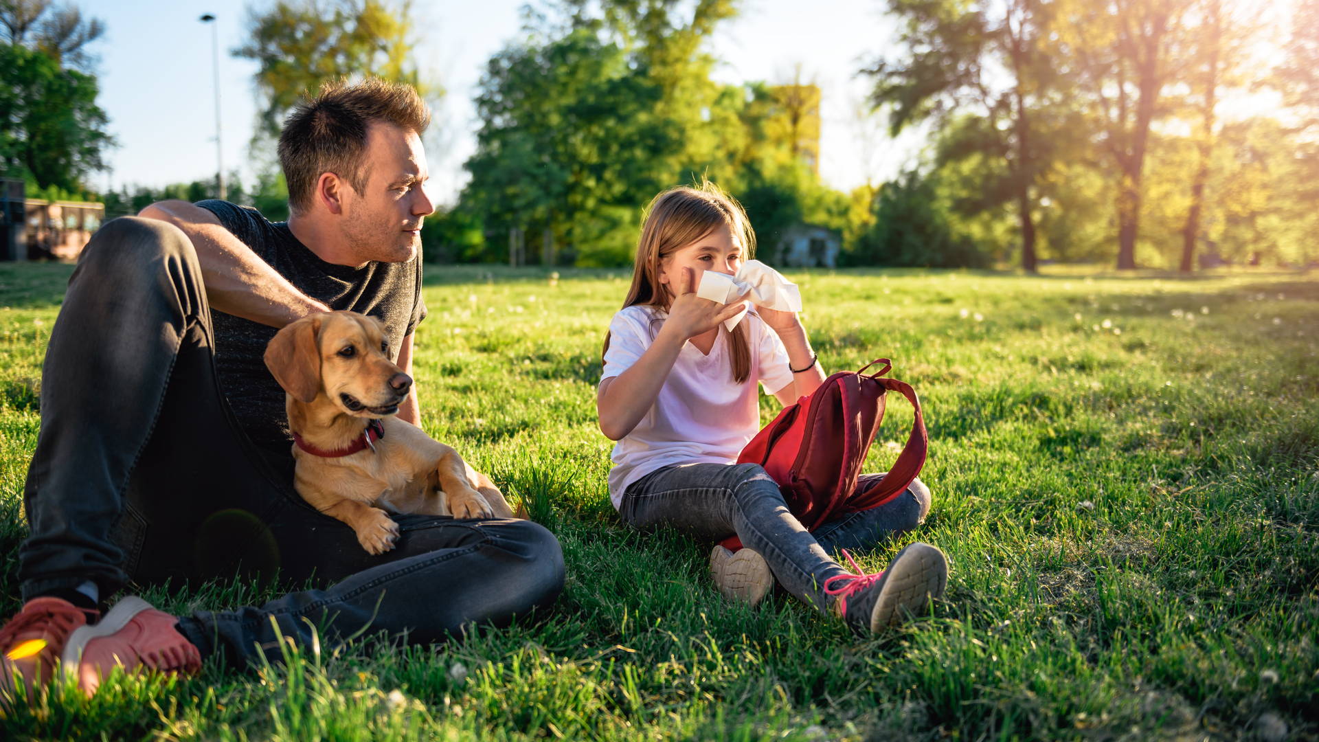 Et barn sitter på en gressplen sammen med en mann og en hund. Barnet tørker nesen med lommetørkle.