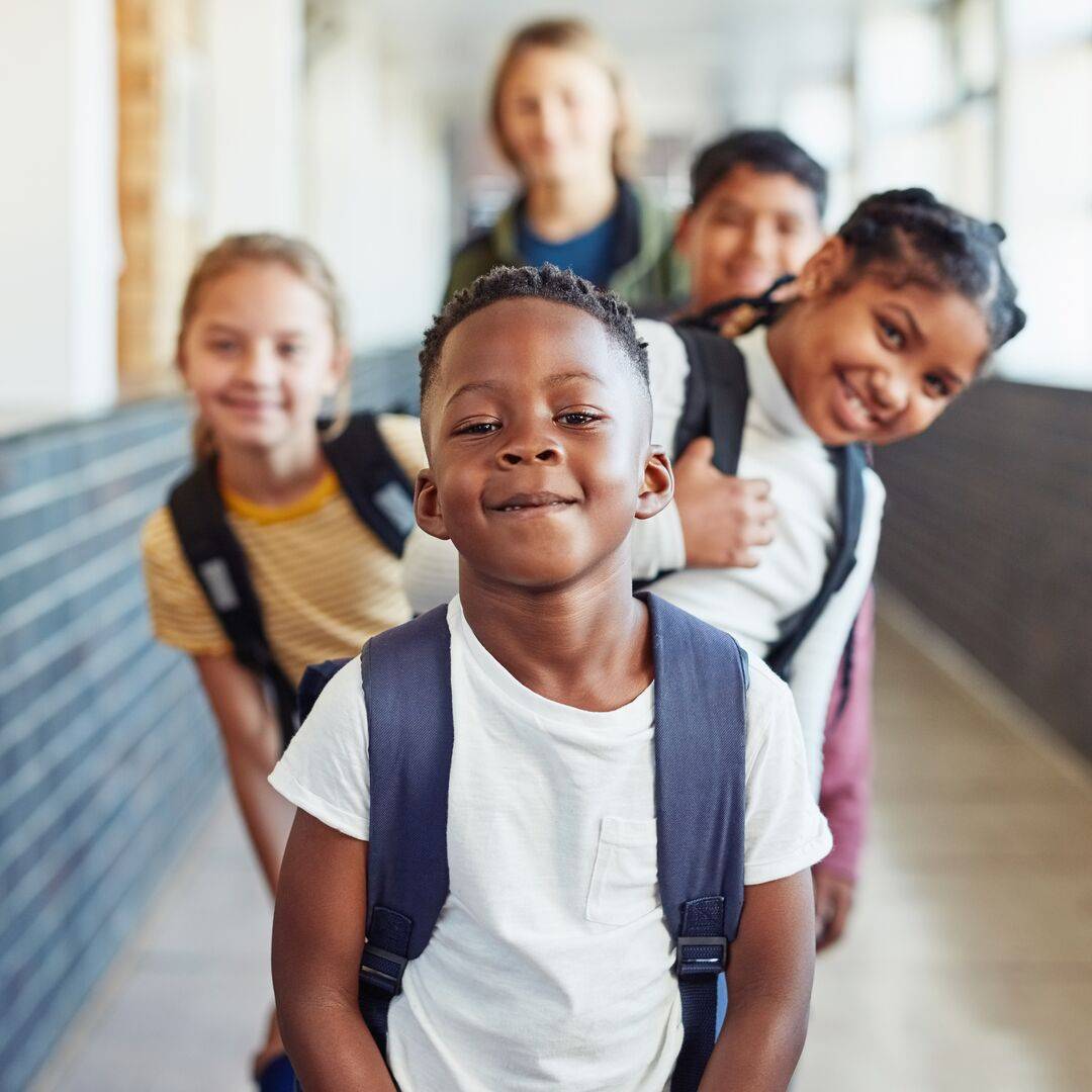 Na školní chodbě jsou před učitelkou seřazeny děti – tři z nich se naklánějí zpoza chlapce vpředu a usmívají se