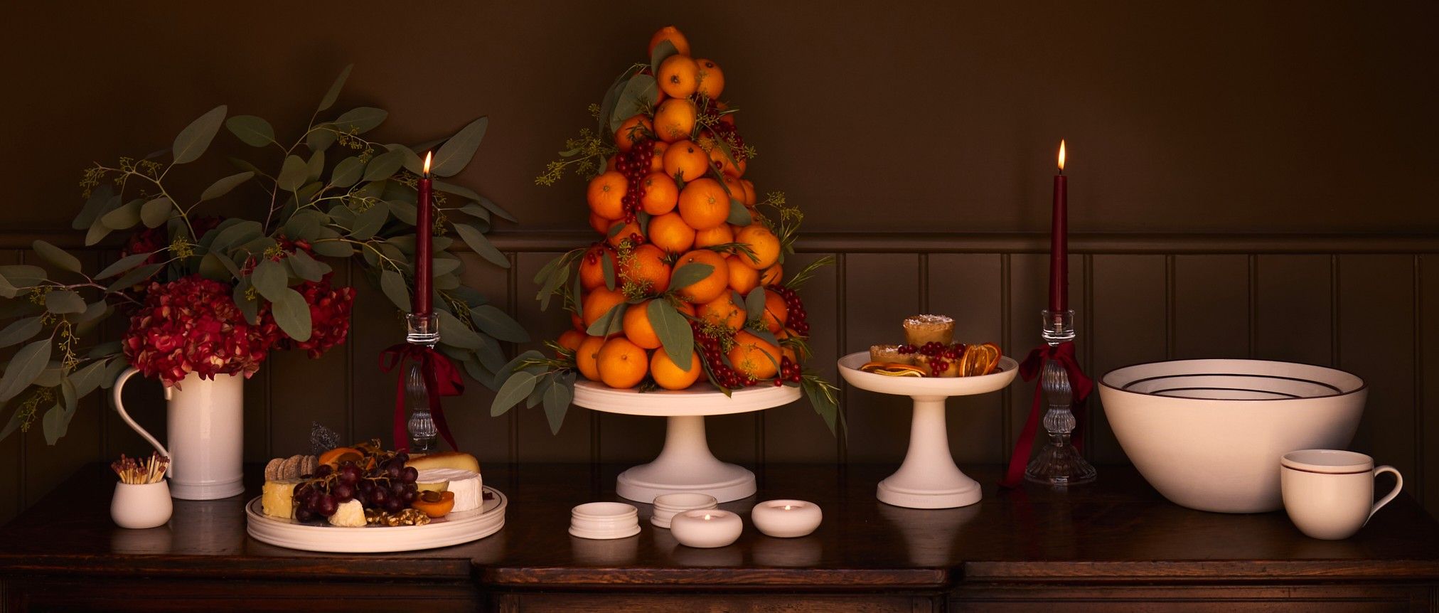 A Festive dresser display of white porcelain ceramics including cake stands and bowls against a dark background. The Cake Stand features a tower of oranges and foliage