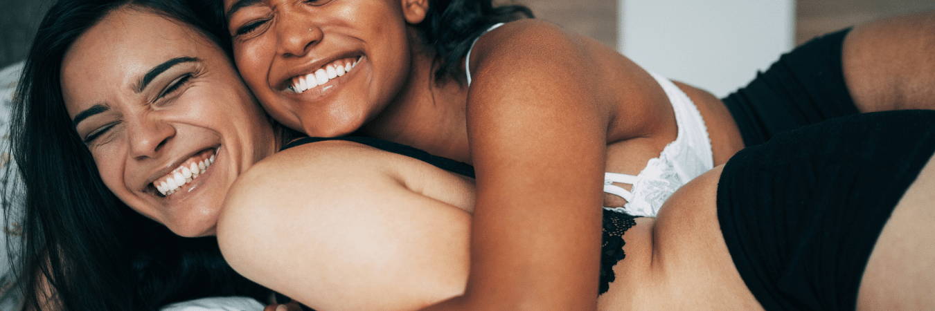 Image of same sex couple in lingerie laughing and smiling on a bed 