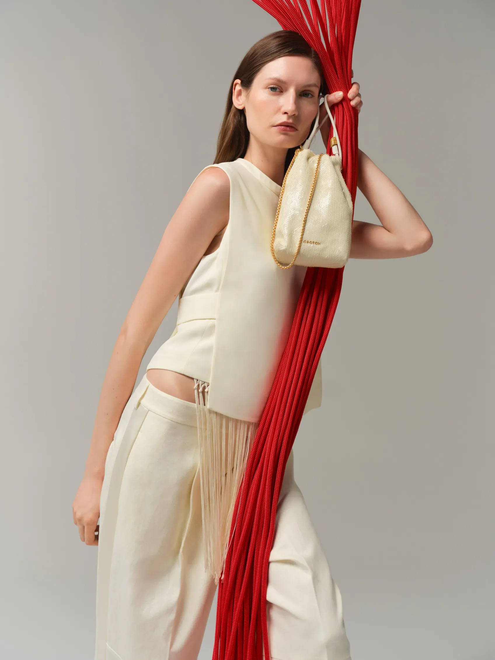 Model in cream outfit holding a bag, with red strands in background.