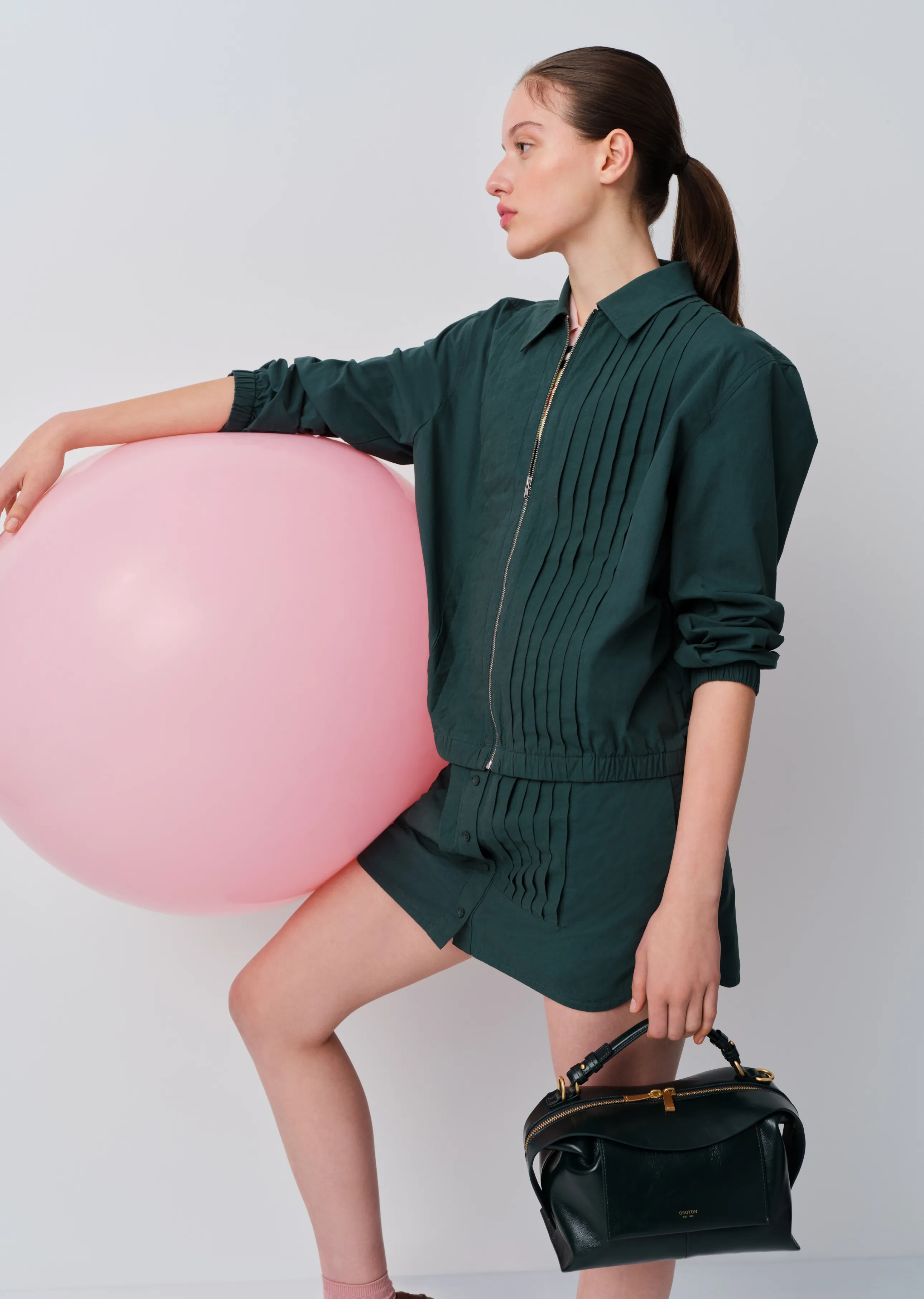 A woman in a green outfit poses with a pink balloon and a handbag.
