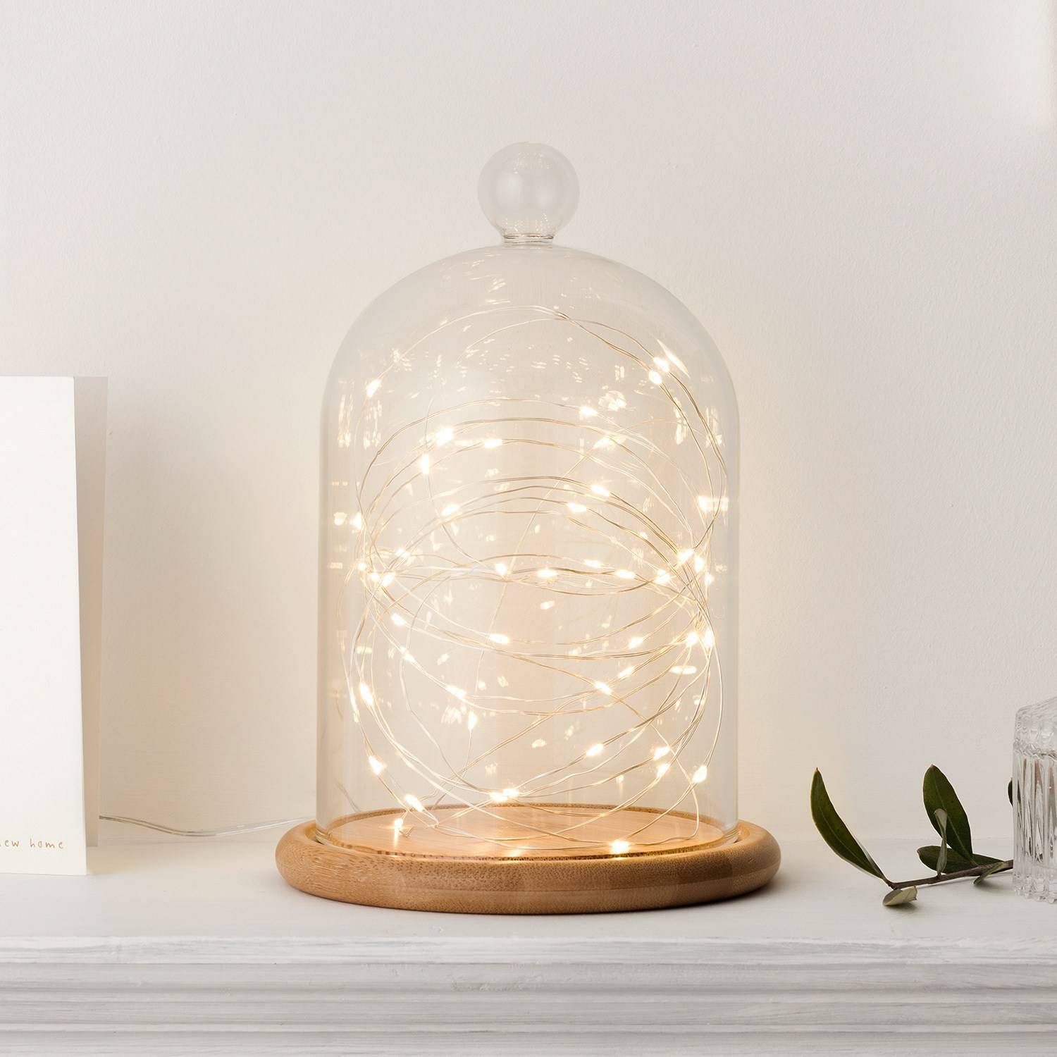 Glass dome with warm white fairy lights inside on a ,amtlepiece with a white wall background.