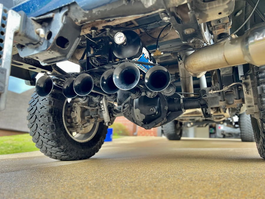 Spare tire delete bay underside Kolyn's blue GMC Sierra Conductor's Special 244K four black Shocker XL horn bells black air tank HornBlasters sticker pressure gauge brass fittings rear axle chrome exhaust off-road tires driveway photo 2
