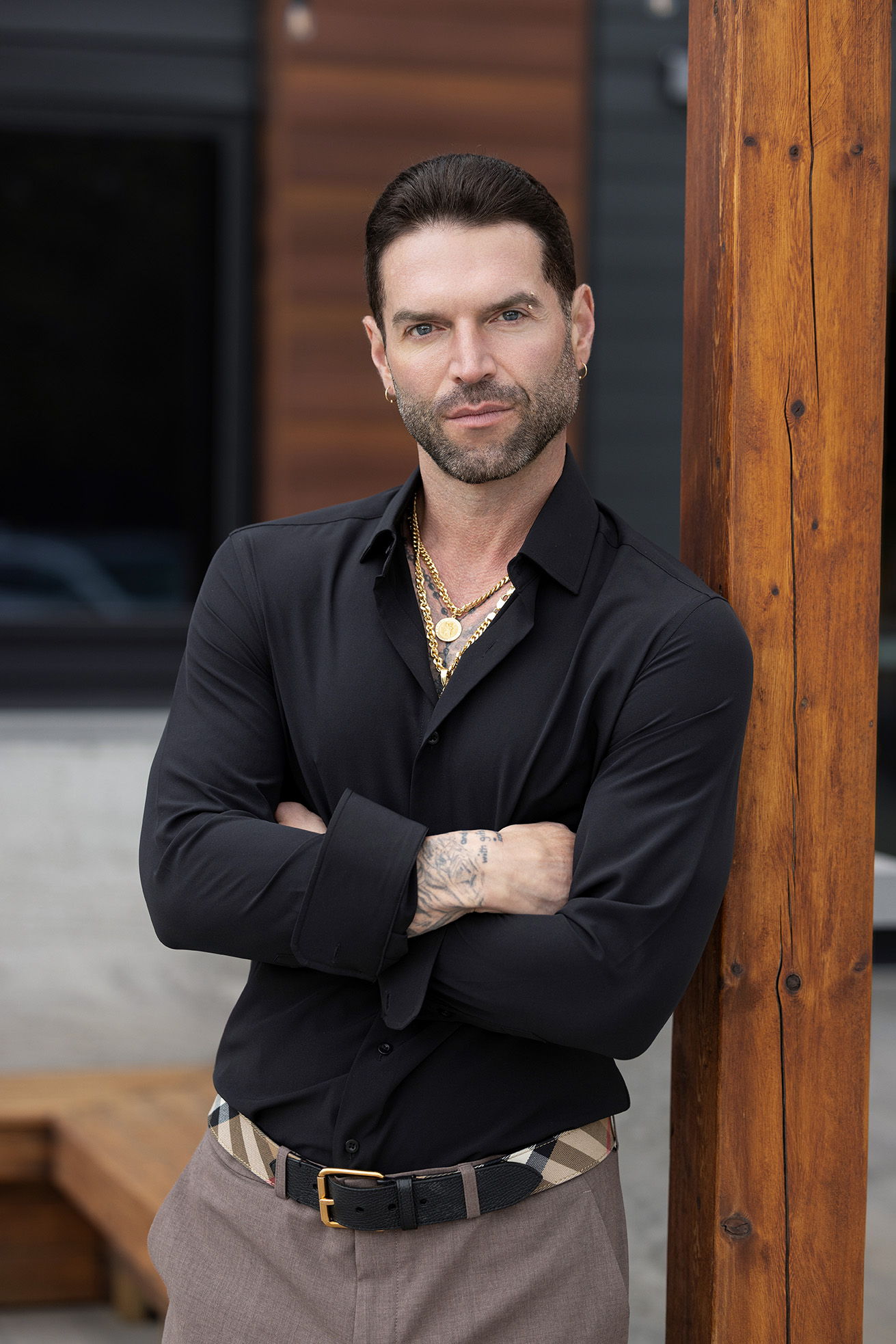 A man in a black shirt poses confidently by a wooden post.