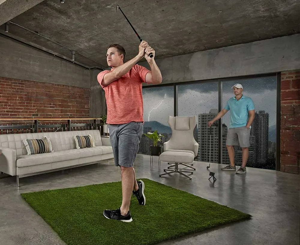 A golfer swinging in a simulator in his apartment using the Garmin Approach R10 while a friend watches