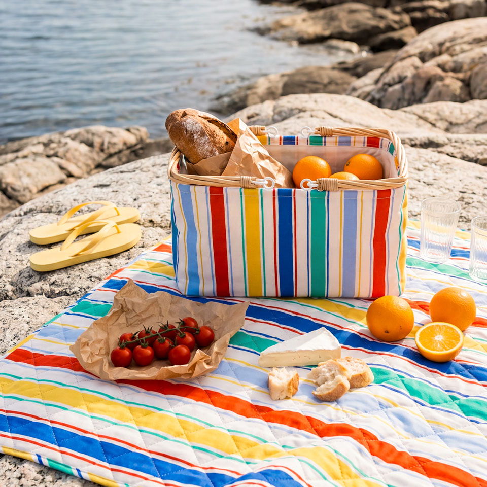 A picnic setup with fruits and bread on a striped blanket by the water.