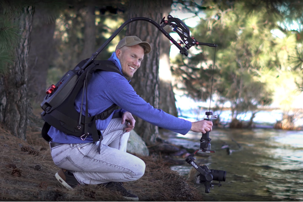 Person crouching by water, holding a camera with a backpack and trees in background.