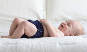 A picture of a baby boy laying on his back on a bed with white sheets and wear a dark blue cloth diaper. 