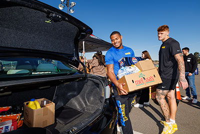 Two individuals carrying food donation boxes near a car in a parking lot.