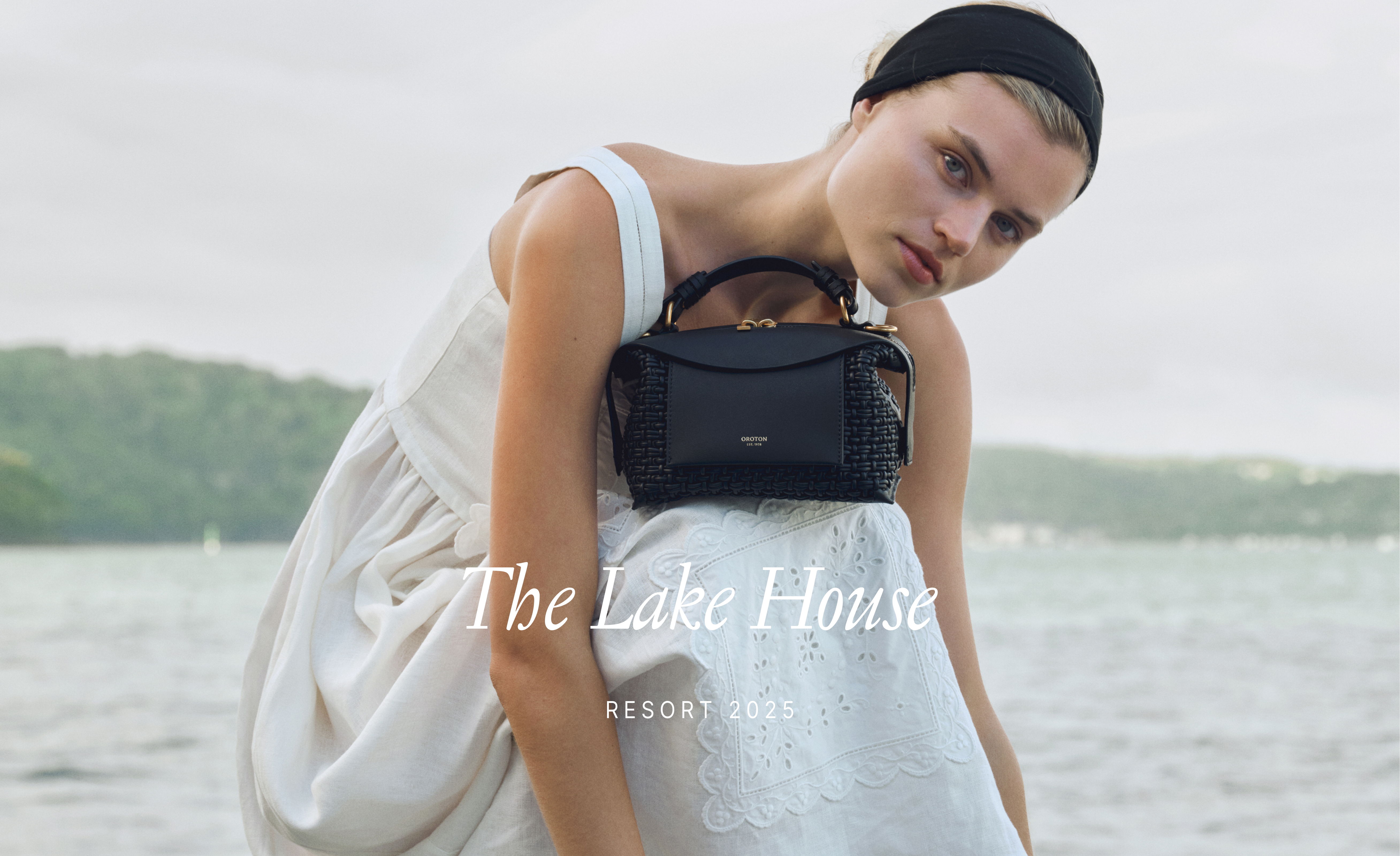 Model wearing a white dress, holding a black handbag by the water.