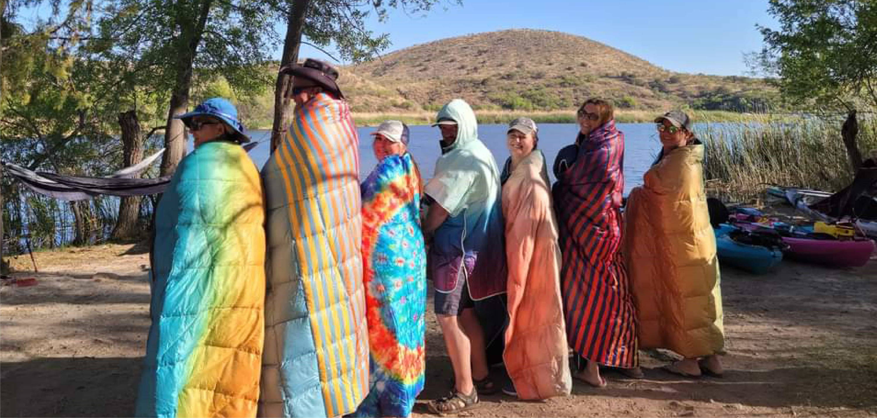 Group Of Men and Womend Wrapped in Rumpl Puffy Blankets On Shore Of River