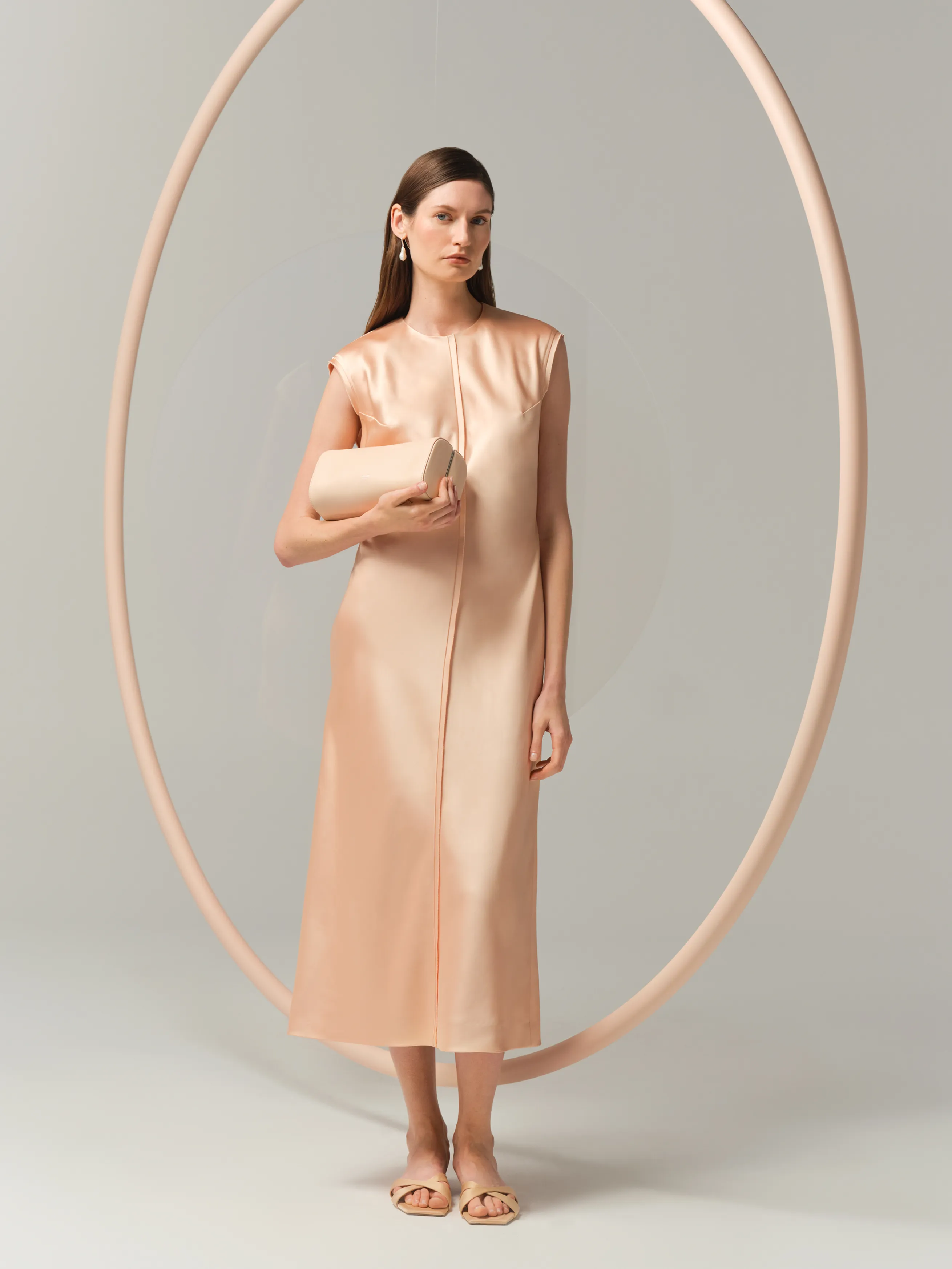 Model in a nude dress holding a clutch, framed by a circular backdrop.