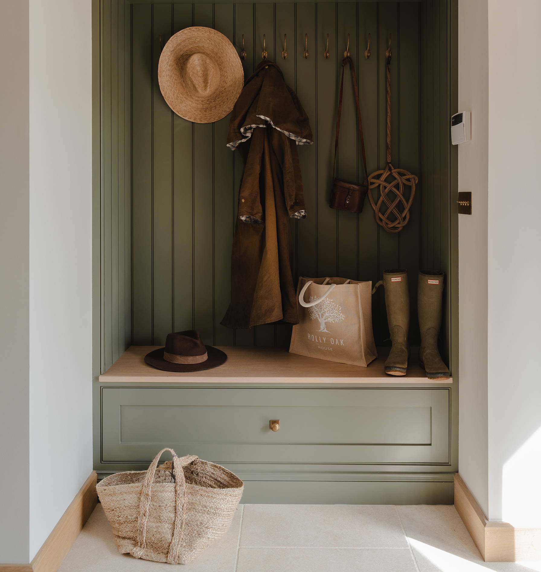 Cosy mudroom design by Holly Oak House with sage green cabinetry, brass hardware, wood panelling, open coat hooks, and tiled flooring