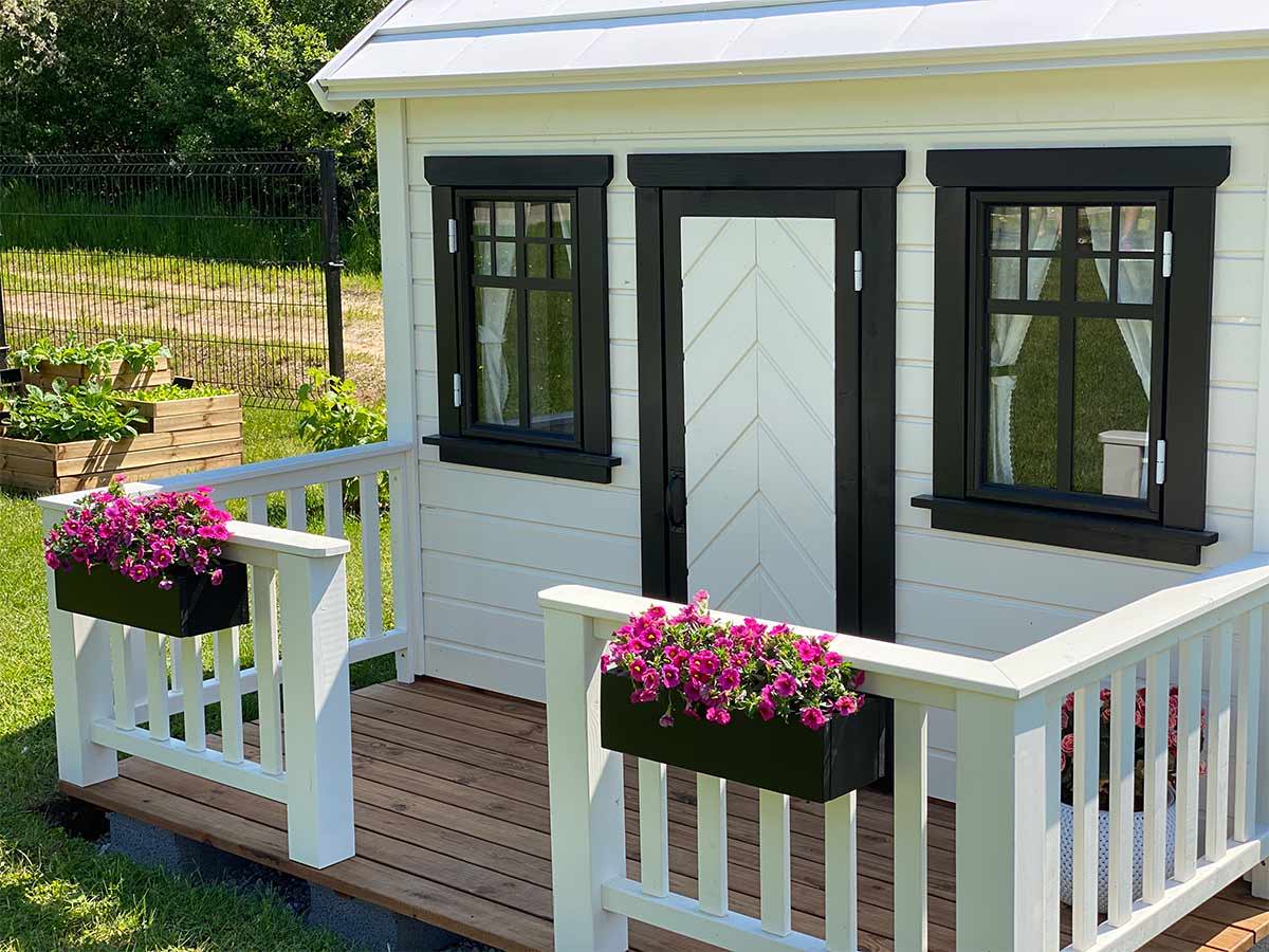 Close up of the front door, windows withs black trims and black flower boxes with pink flowers of  Custom Wooden Playhouse by WholeWoodPlayhouses