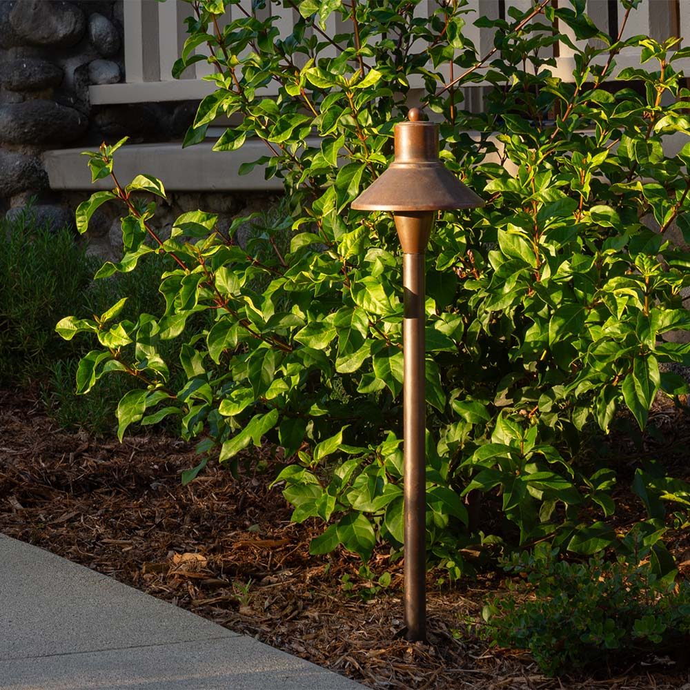 A brightly sunlit flared-top pathway light sits in some brush in front of a porch.