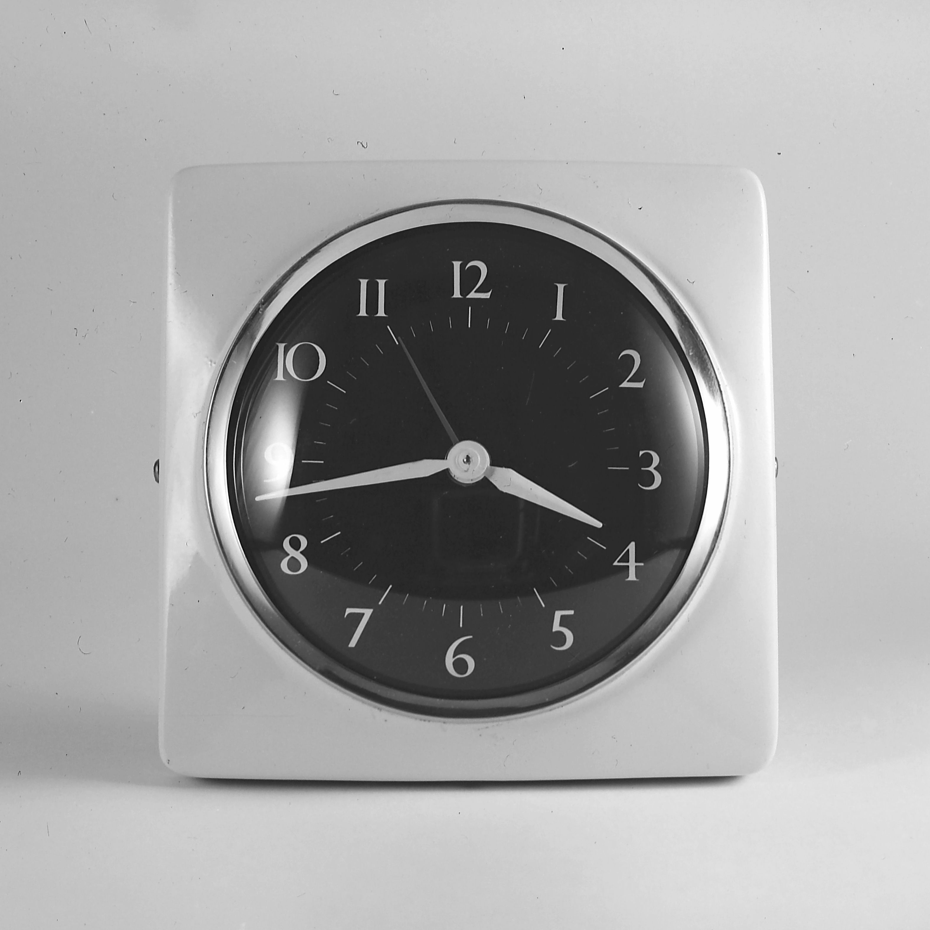 A square clock with black face and white numbers on a gray background.
