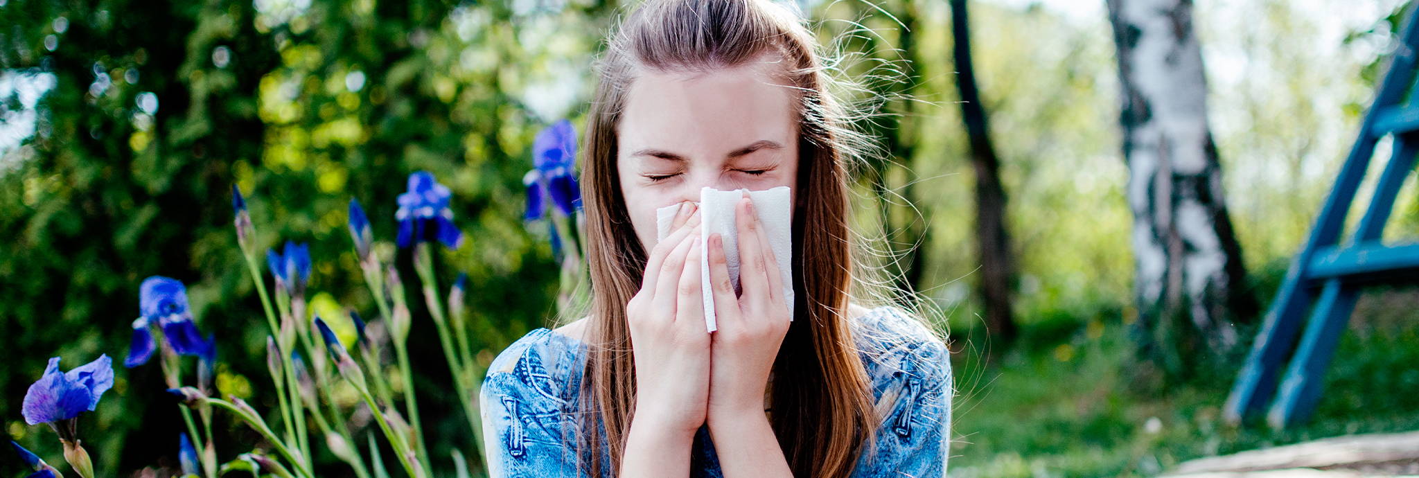 Eine Jugendliche putzt sich ihre laufende Nase – Sie sitzt an einem sonnigen Tag draußen und hinter ihr sind Birken. Vielleicht hat sie eine Birkenpollenallergie.