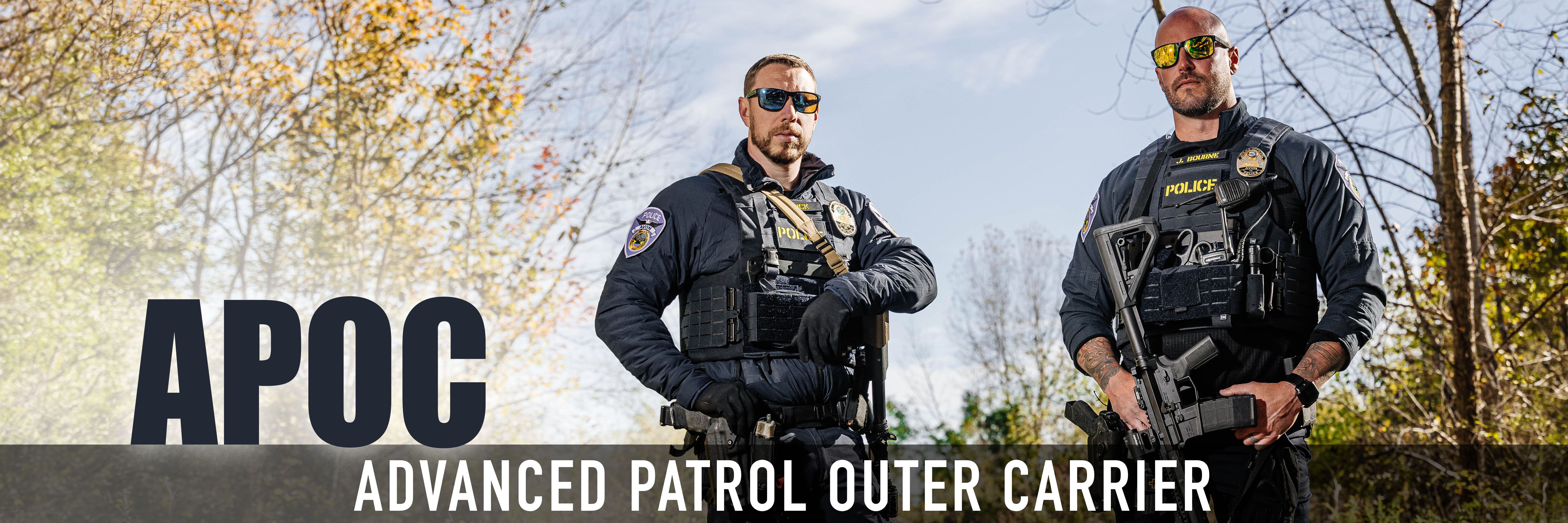 Two police officers in tactical gear standing outdoors with trees in background.