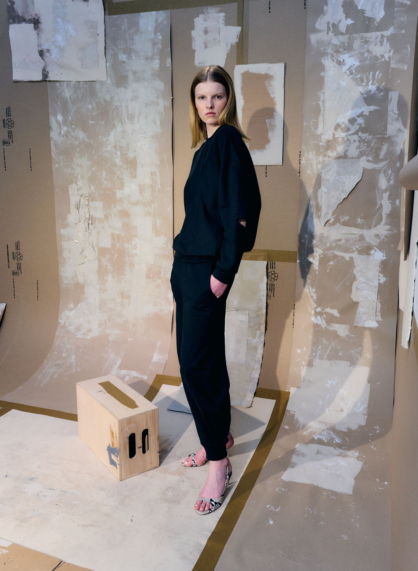 Model in black outfit standing on a textured floor with cardboard backdrop.