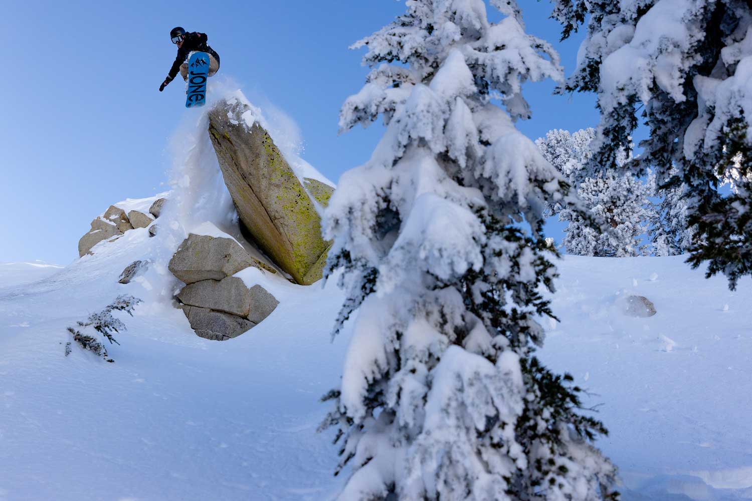 Person jumping off a snow-covered rock with trees in the background.
