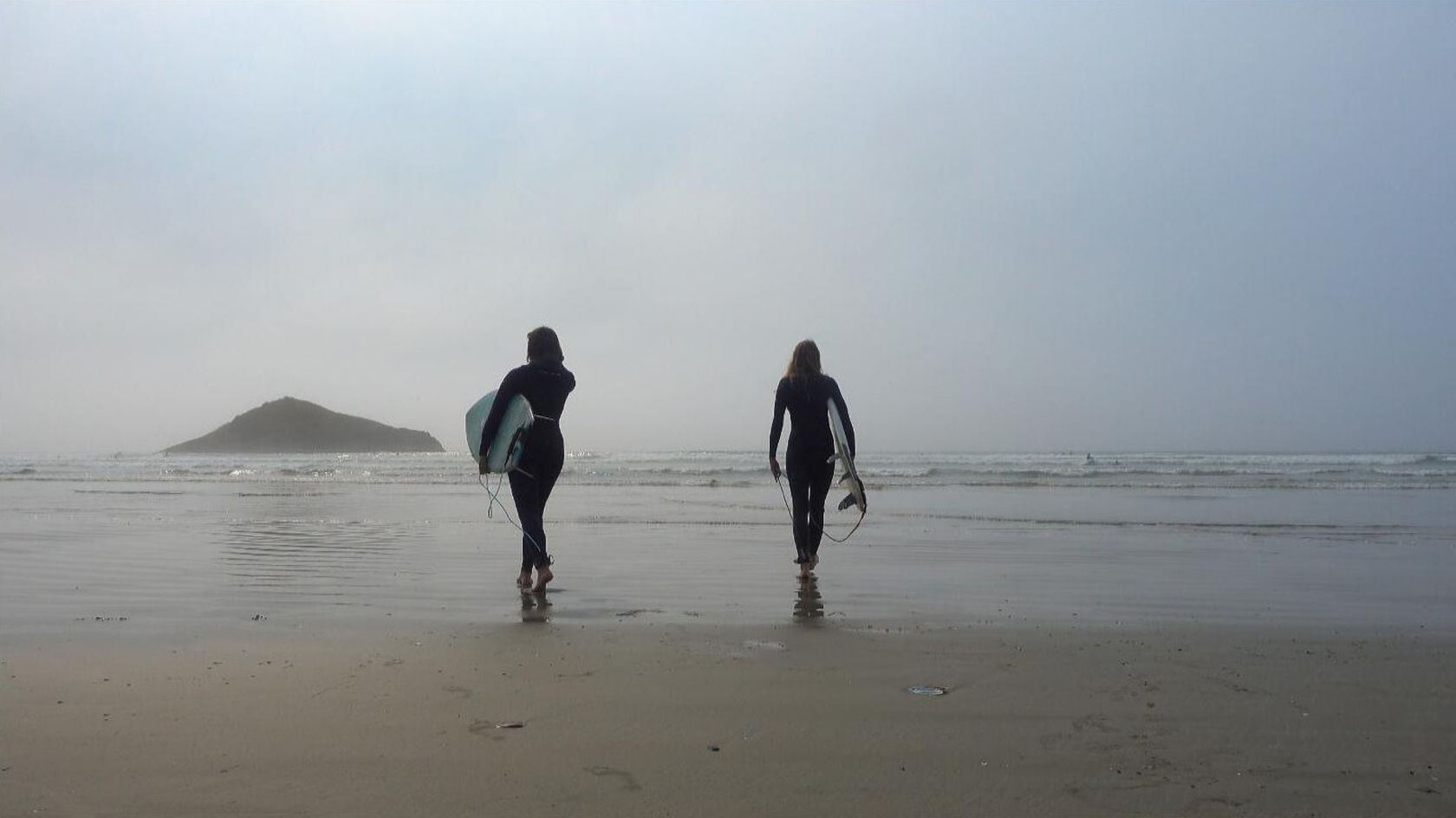 Two surfers walking along a foggy beach beside the ocean.