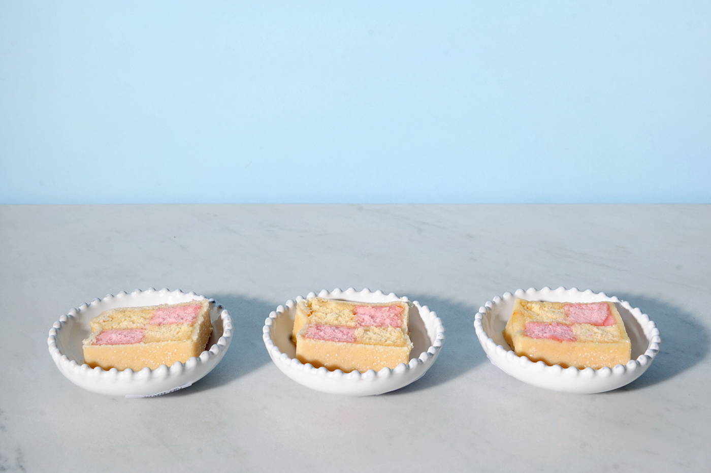 A trio of Costa Nova Pearl White mini bowls in a line on a marble table. Each bowl contains a slice of Battenburg cake.