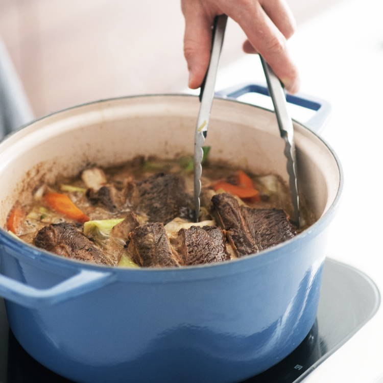  A blue Misen Dutch Oven filled with braised meat and vegetables, with a hand holding tongs reaching inside.
