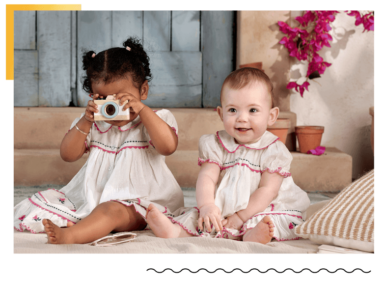 Two babies sitting together, one holding a camera, smiling.