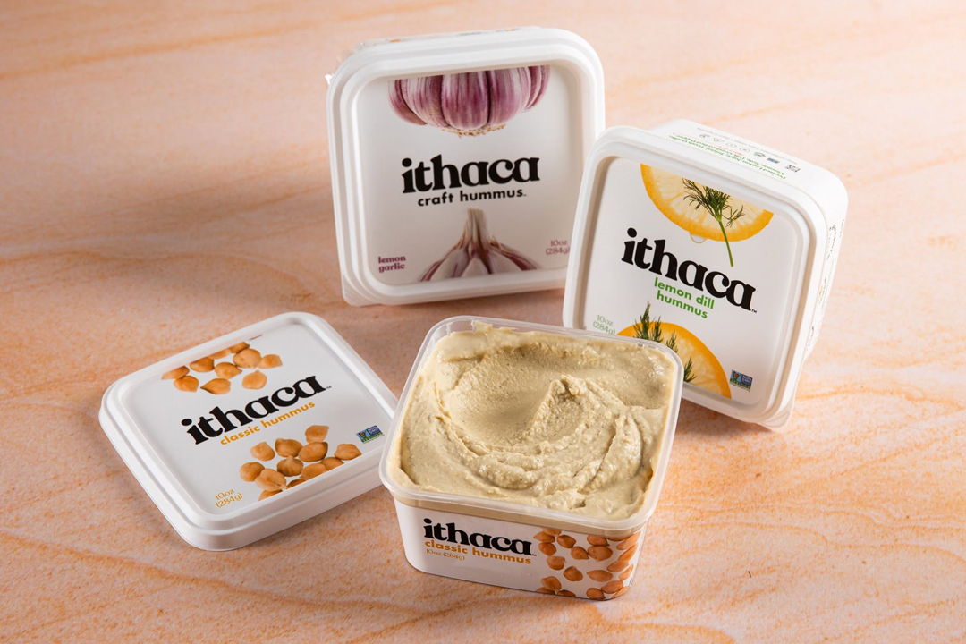 Four containers of Ithaca hummus with different flavors on a wooden surface.
