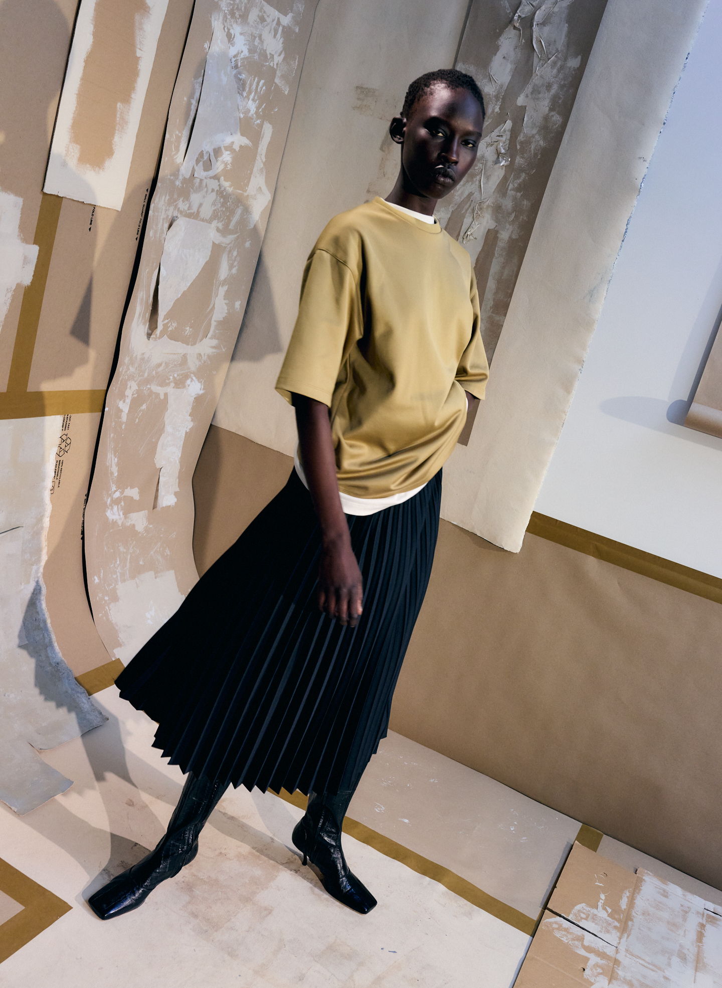 Model wearing a yellow shirt and black pleated skirt in an artistic setting.