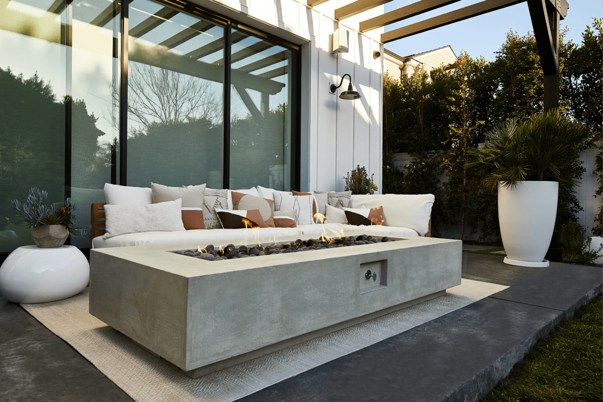 A concrete fire table and plush sofa on a patio under a wooden pergola. 