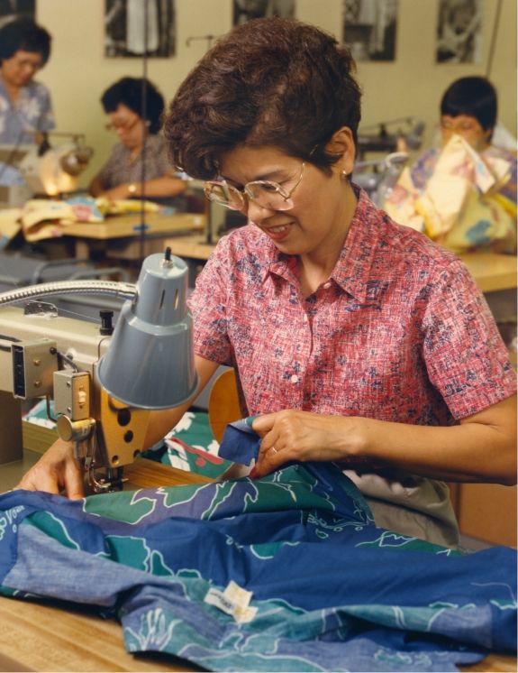 Woman sewing fabric at a table in a workshop setting.
