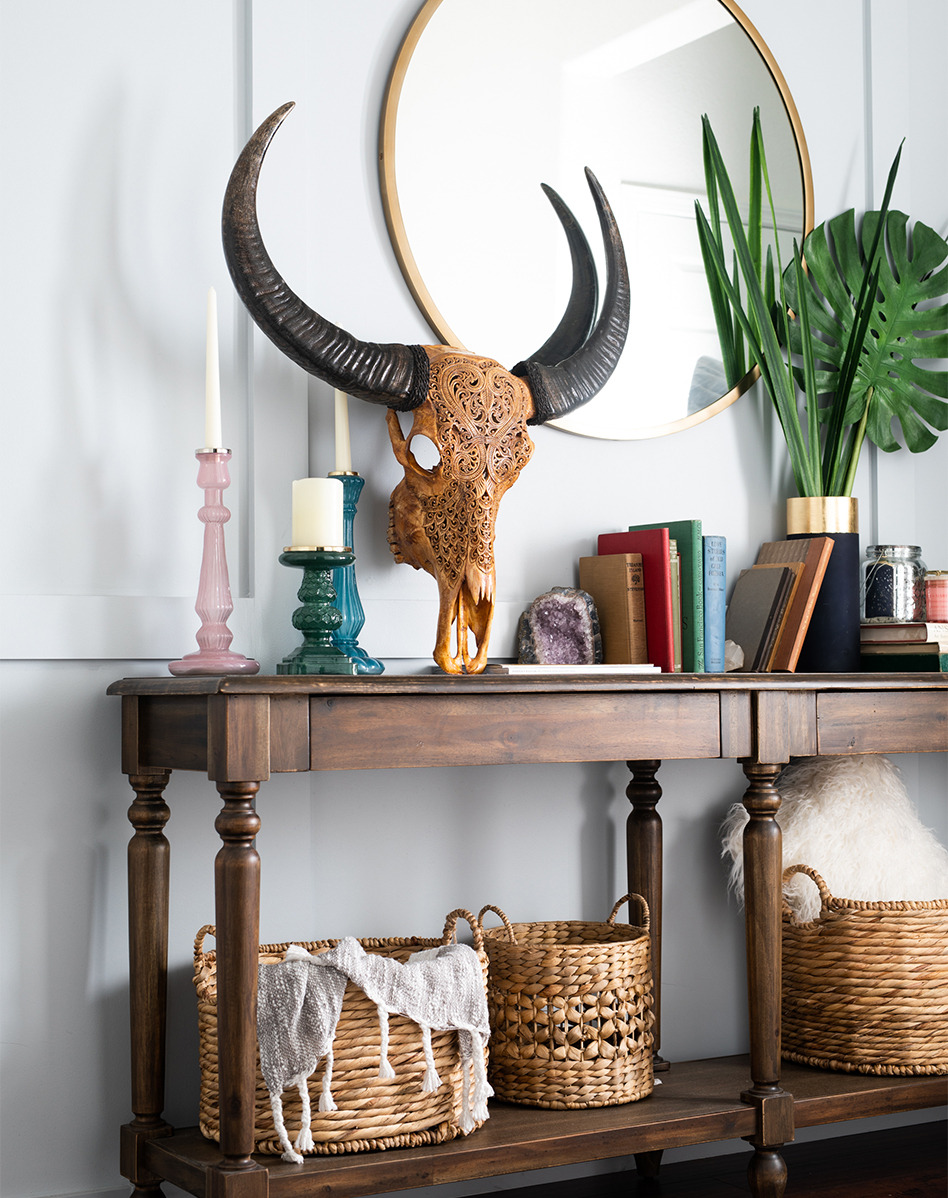 A hand carved skull, books, candles, plants and baskets are presented on a rustic shelf.