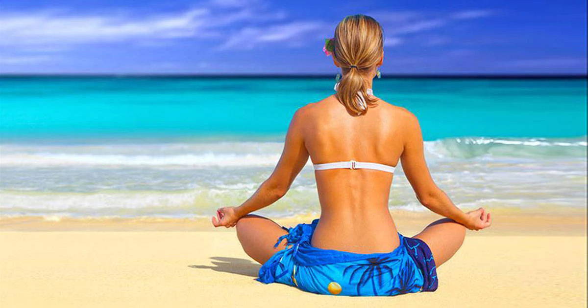 white woman wearing a white bikini halter top and blue sarong, sitting on the beach in cross-legged mediation pose, facing the ocean