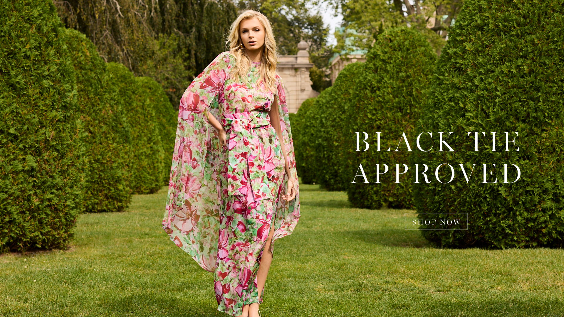 Model in floral dress standing in garden with hedges.