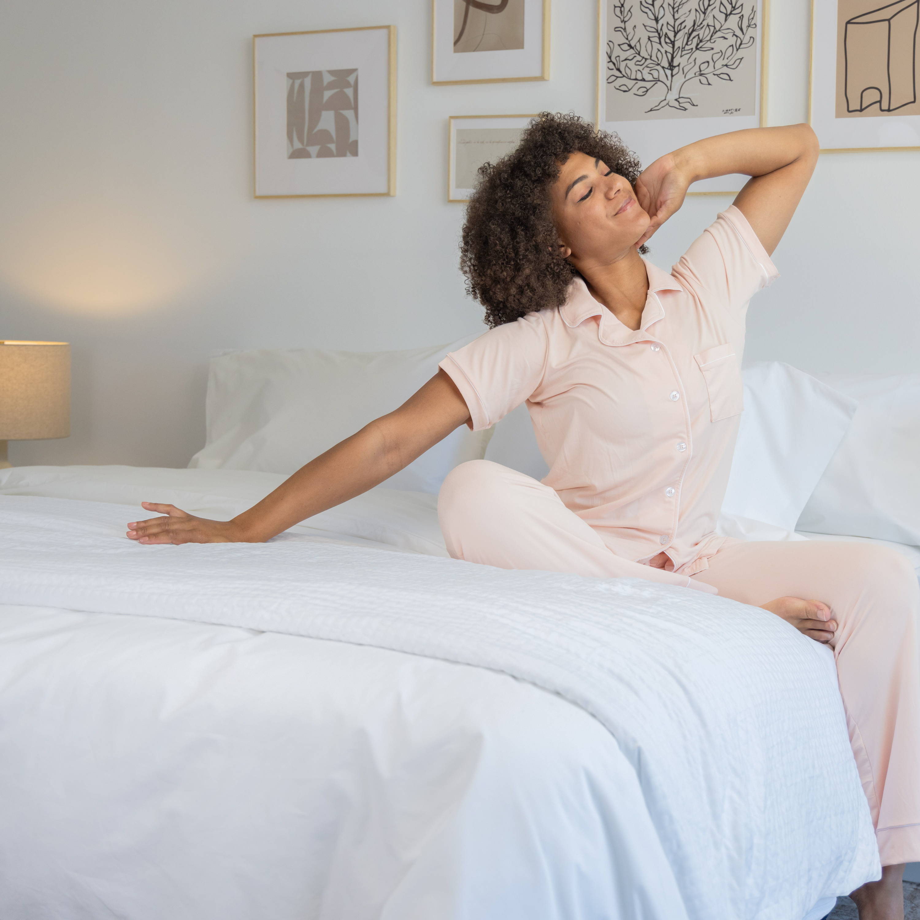 Woman stretching on the edge of her bed, white Comfortcore and Percale bedding, with wooden night tables and ceramic blue lamps