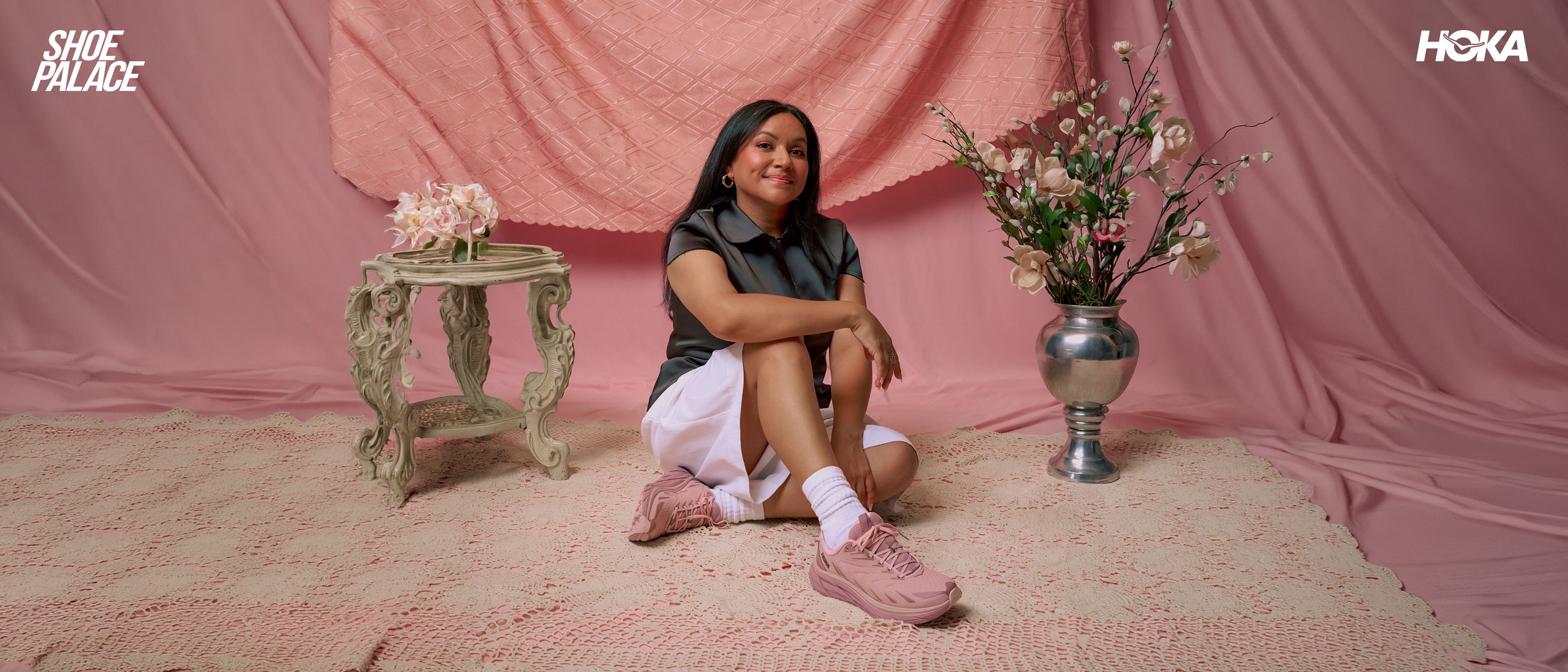 Young woman sitting on floor, surrounded by pink backdrop and flowers.