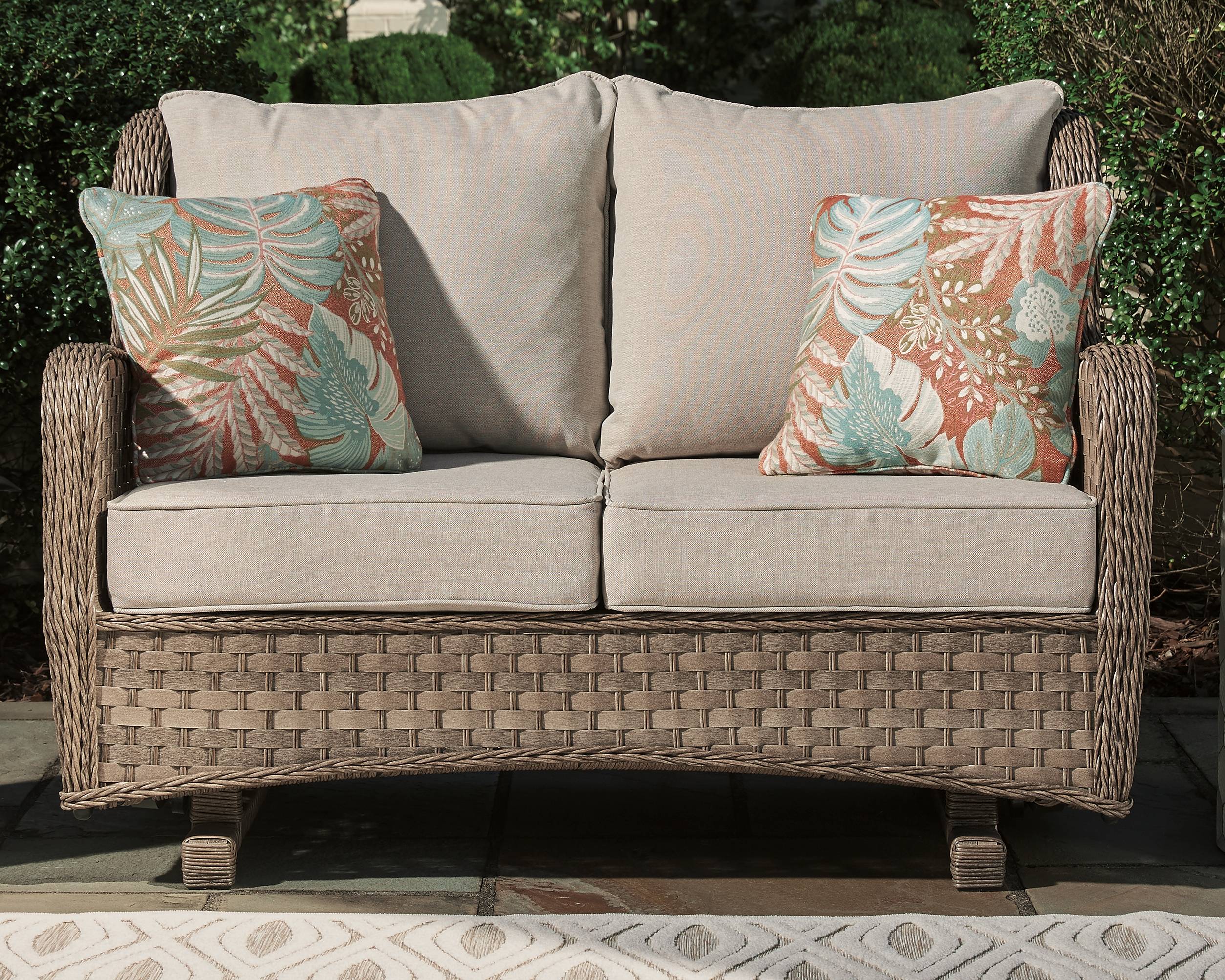 A woven wicker outdoor loveseat sits on patio in front of a wall of greenery. Two grey cushions support two orange floral outdoor pillows on either side of the sofa.