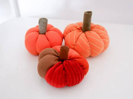 Three finished fabric pumpkins made out of assorted orange fabric scraps on a white table