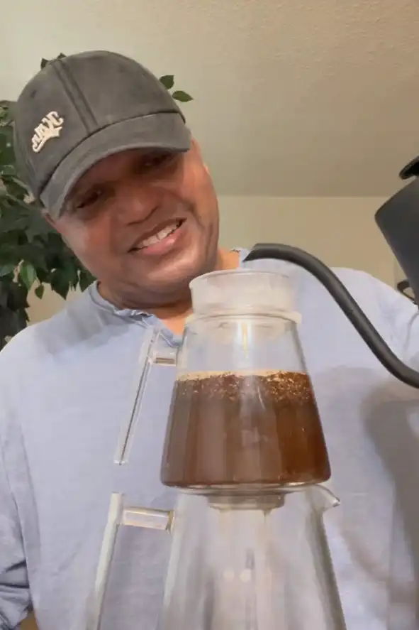 Person brewing coffee with a coffee maker, smiling and wearing a cap.