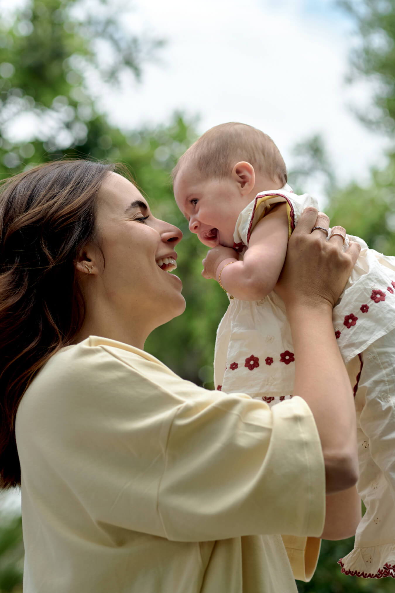 A mother joyfully holds and plays with her smiling baby outdoors.