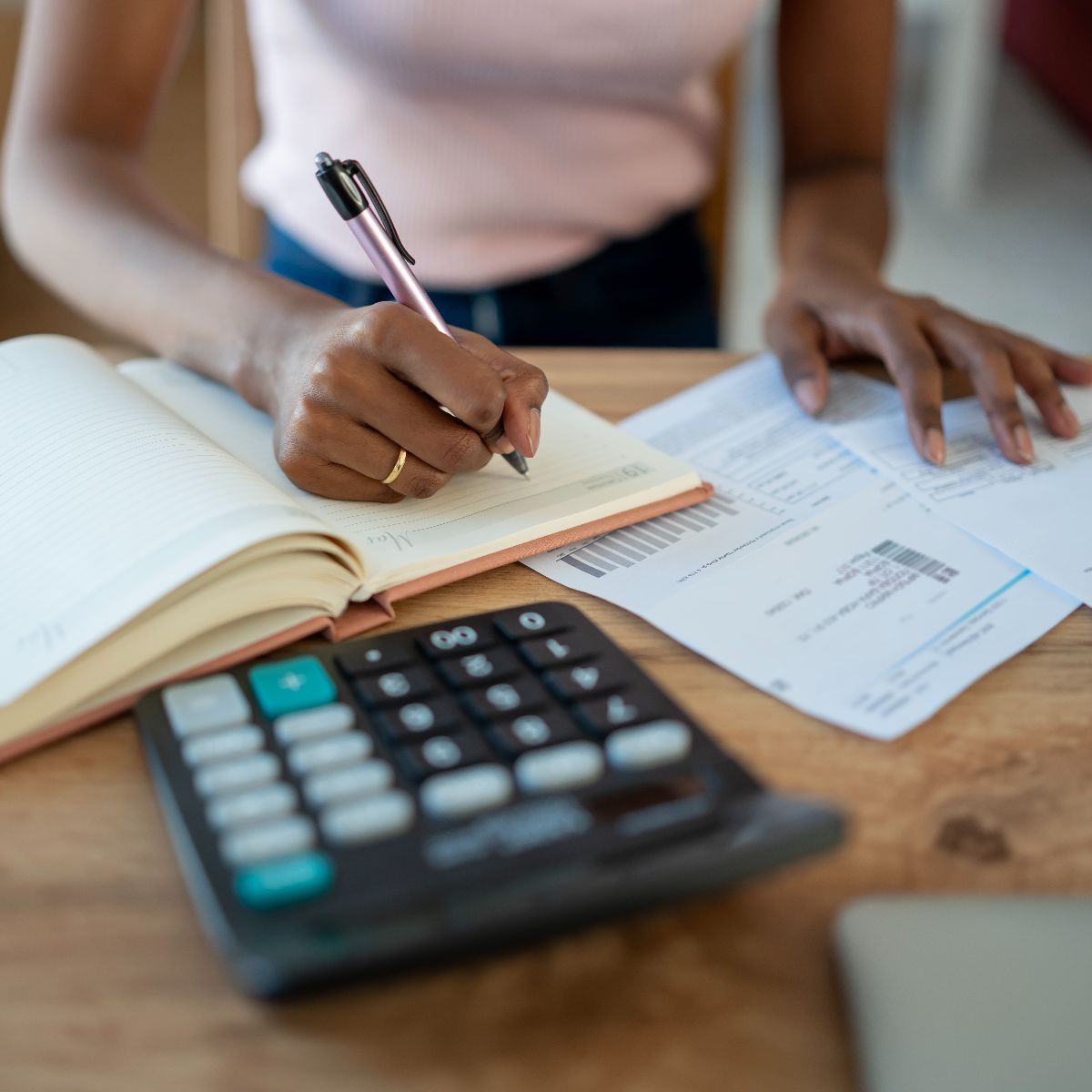 Person writing in a notebook with bills and a calculator on a desk.