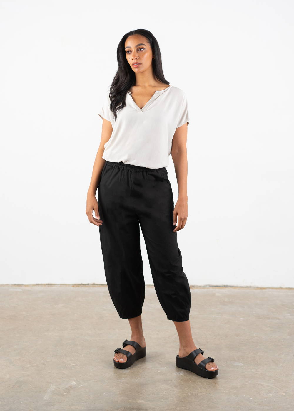 A model wearing a white v-neck short sleeved top with black balloon leg tapered trousers with black chunky platform slides
