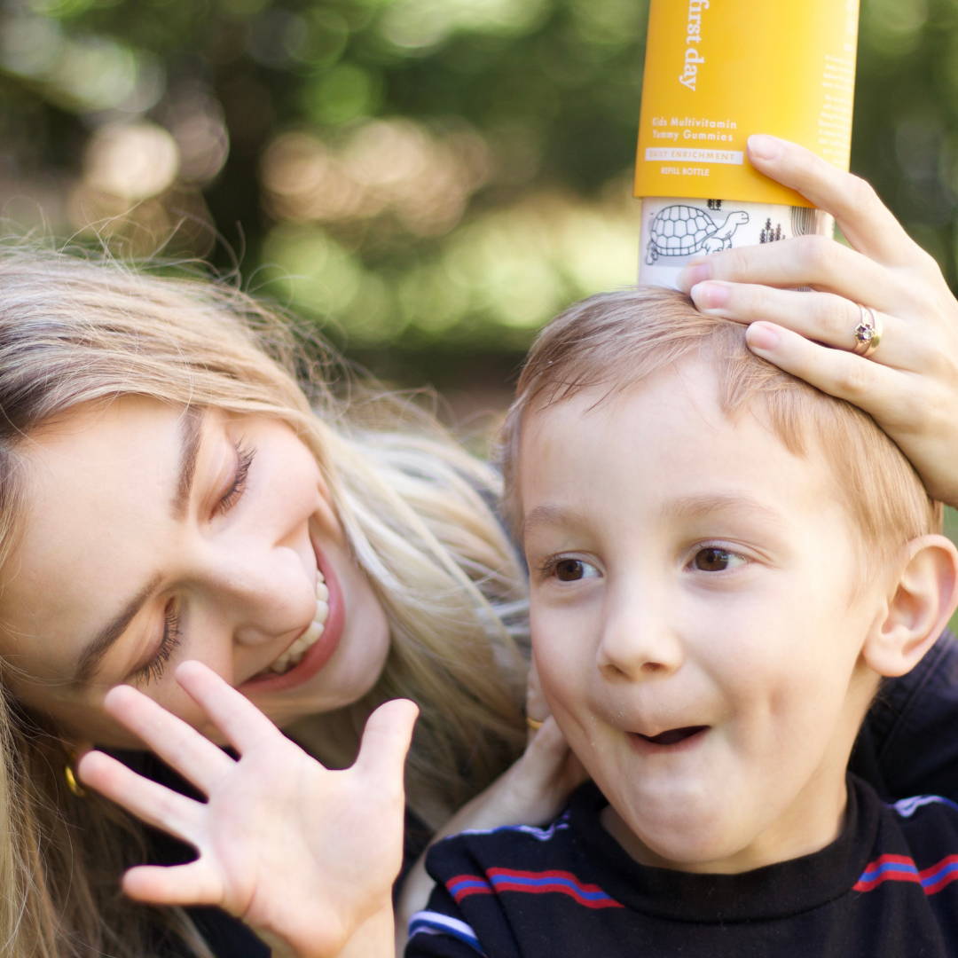 A mom playing with her son, holding the vitamin bottle on his head while he smiles