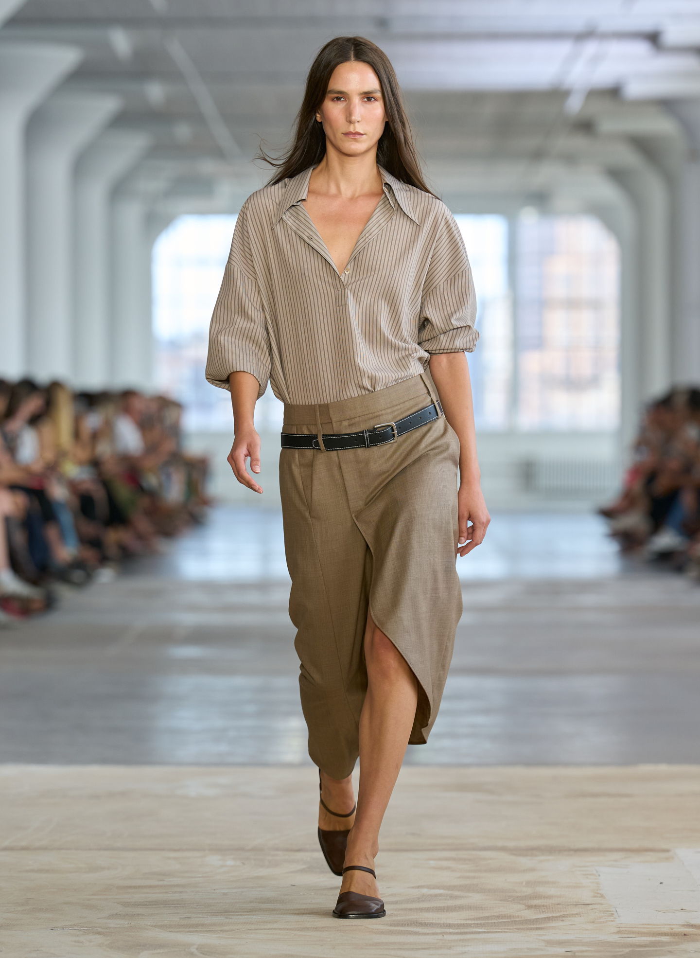 Model walking down runway in neutral-colored outfit during a fashion show.