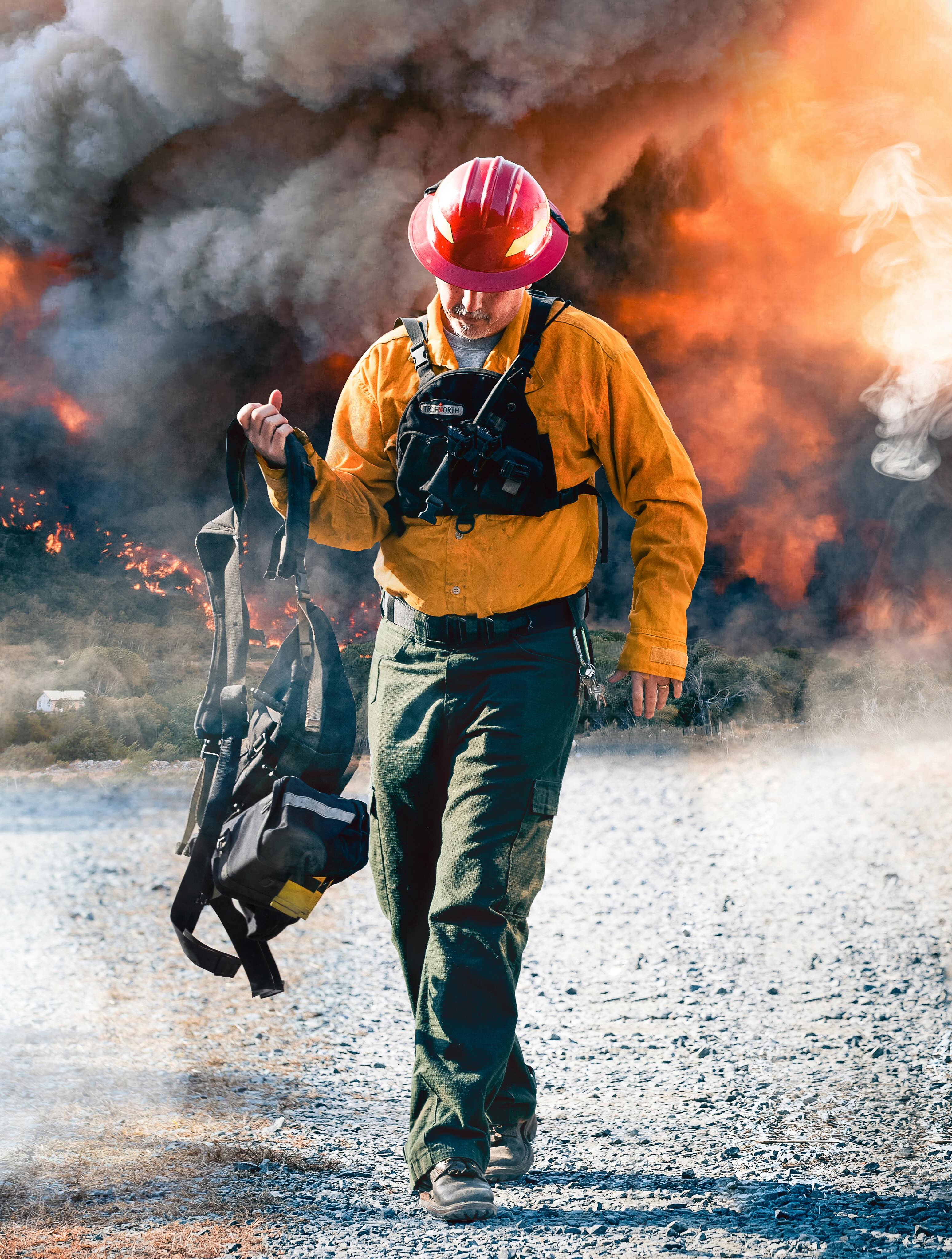 Firefighter walking away from flames, carrying equipment in a smoky environment.