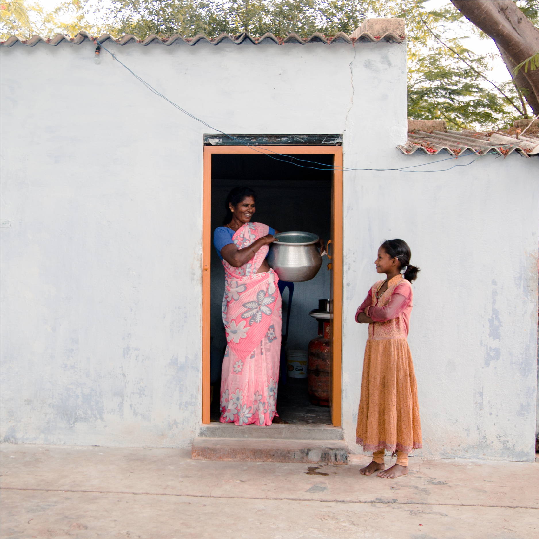 An Indian woman stands next to a young girl in a doorway holding a pot of food. 